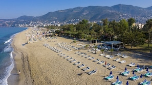 A beach with rows of sun loungers and umbrellas, a lifeguard tower, and a backdrop of mountains and a city. - Aslan City Hotel (Alanya)