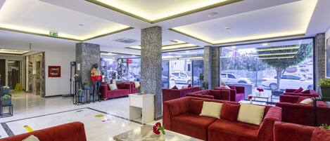 A hotel lobby with red sofas, a marble coffee table, and large windows offering a view of parked cars outside.