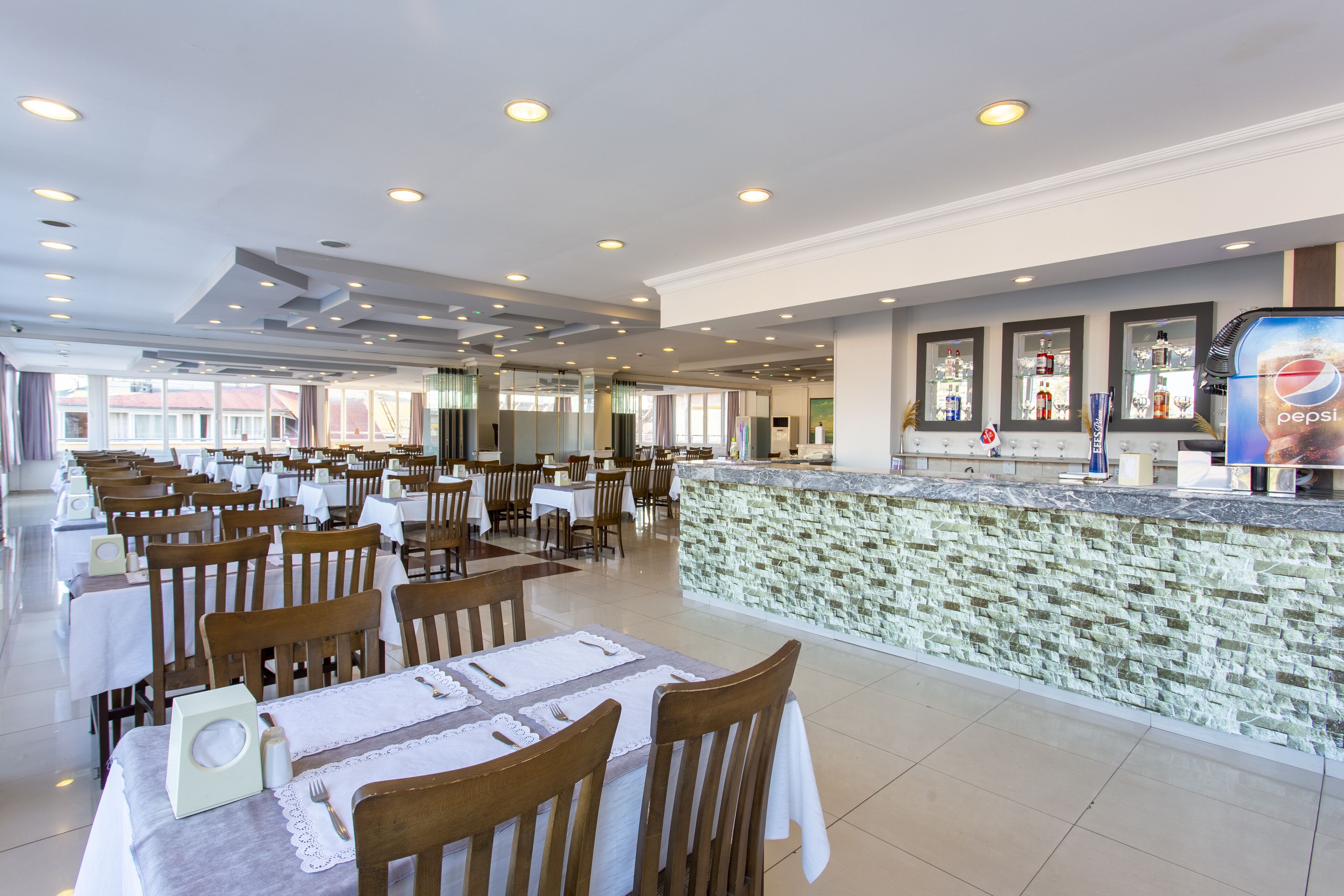 A spacious restaurant with multiple tables and chairs, a stone counter with a Pepsi advertisement, and a view of the outside.
