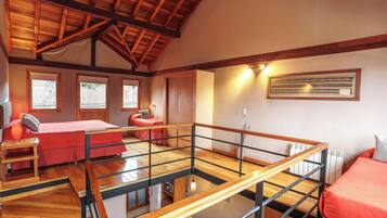 A spacious room with a high ceiling, wooden beams, a bed with a red bedspread, a desk with a lamp, and a framed picture on the wall.
