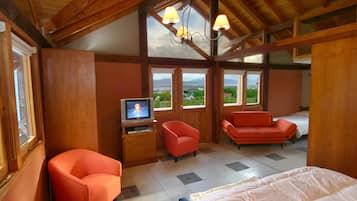 Quarto com cama, duas cadeiras vermelhas, televisão e janelas amplas com vista para o exterior.