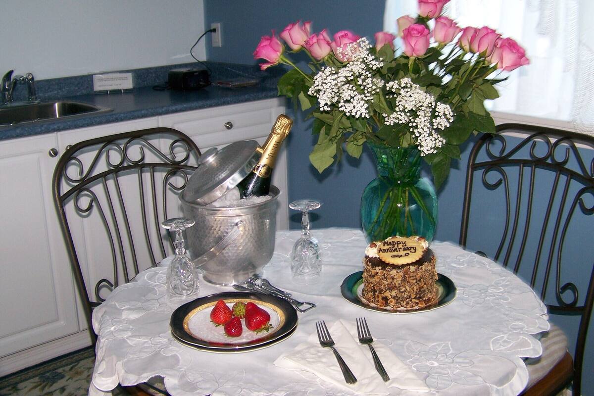 Uma mesa posta com bolo, morangos, uma garrafa de champanhe e um vaso de rosas cor-de-rosa.
