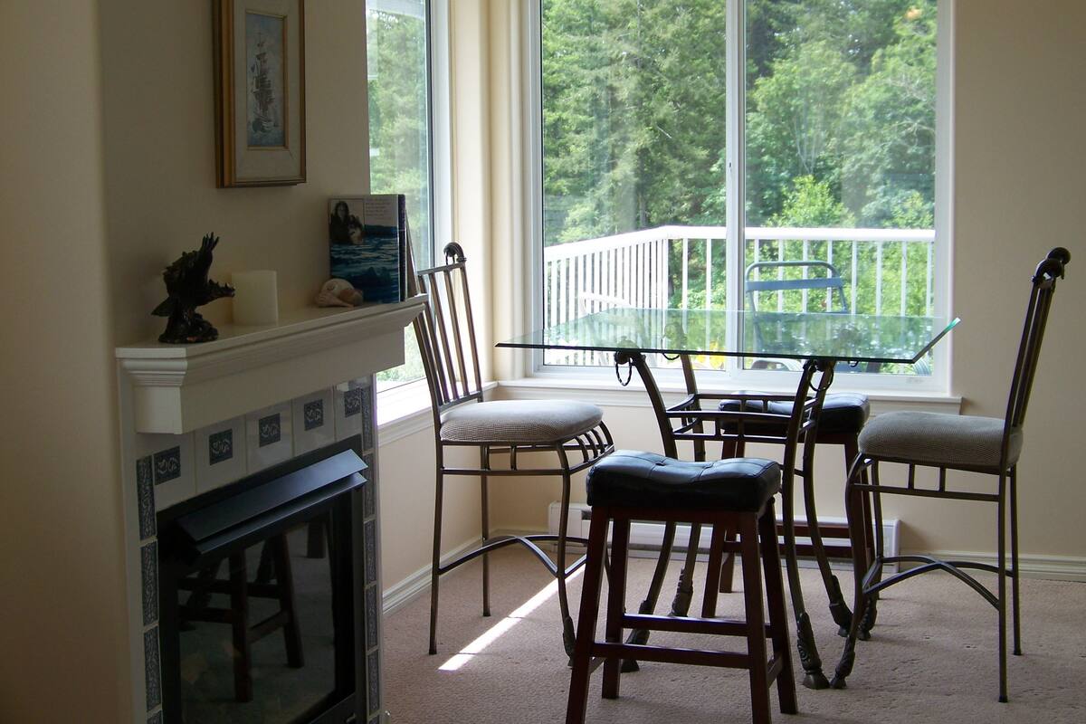 Uma sala de jantar com mesa e cadeiras de vidro, lareira e vista para uma varanda com guarda-corpo.
