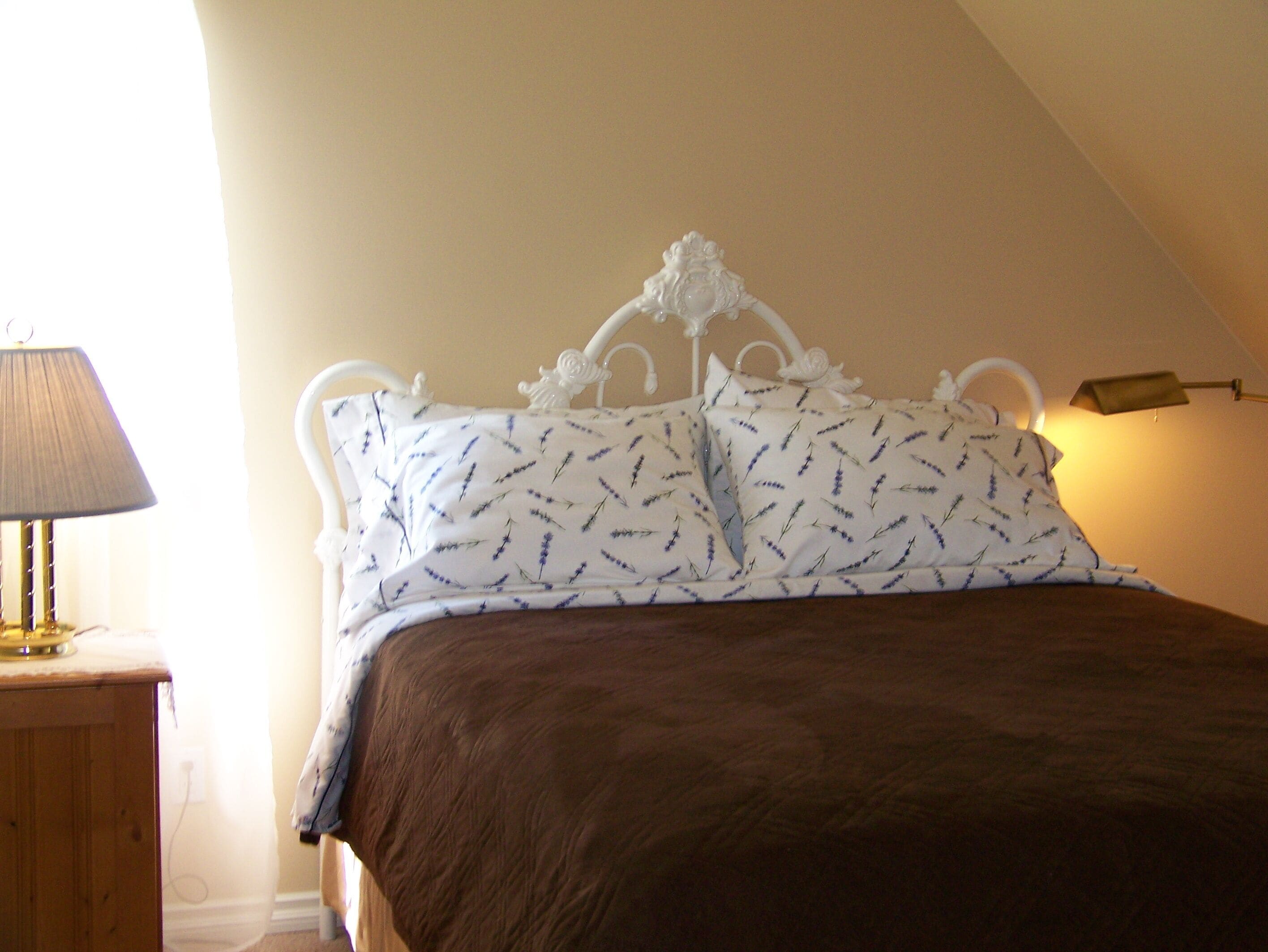 A bedroom with a white ornate headboard, lavender-patterned pillows, a brown bedspread, and a bedside lamp.