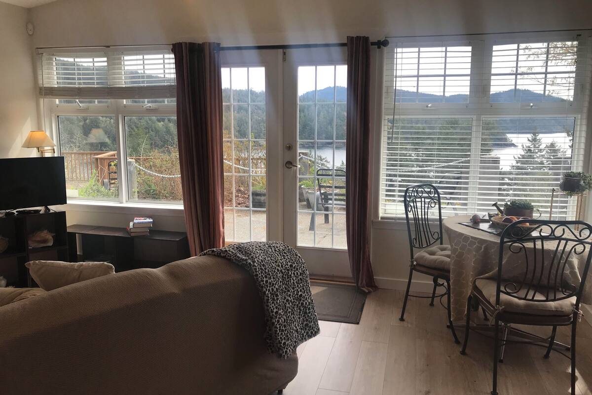 Uma sala de estar com sofá, televisão, mesa e cadeiras, e amplas janelas com cortinas.