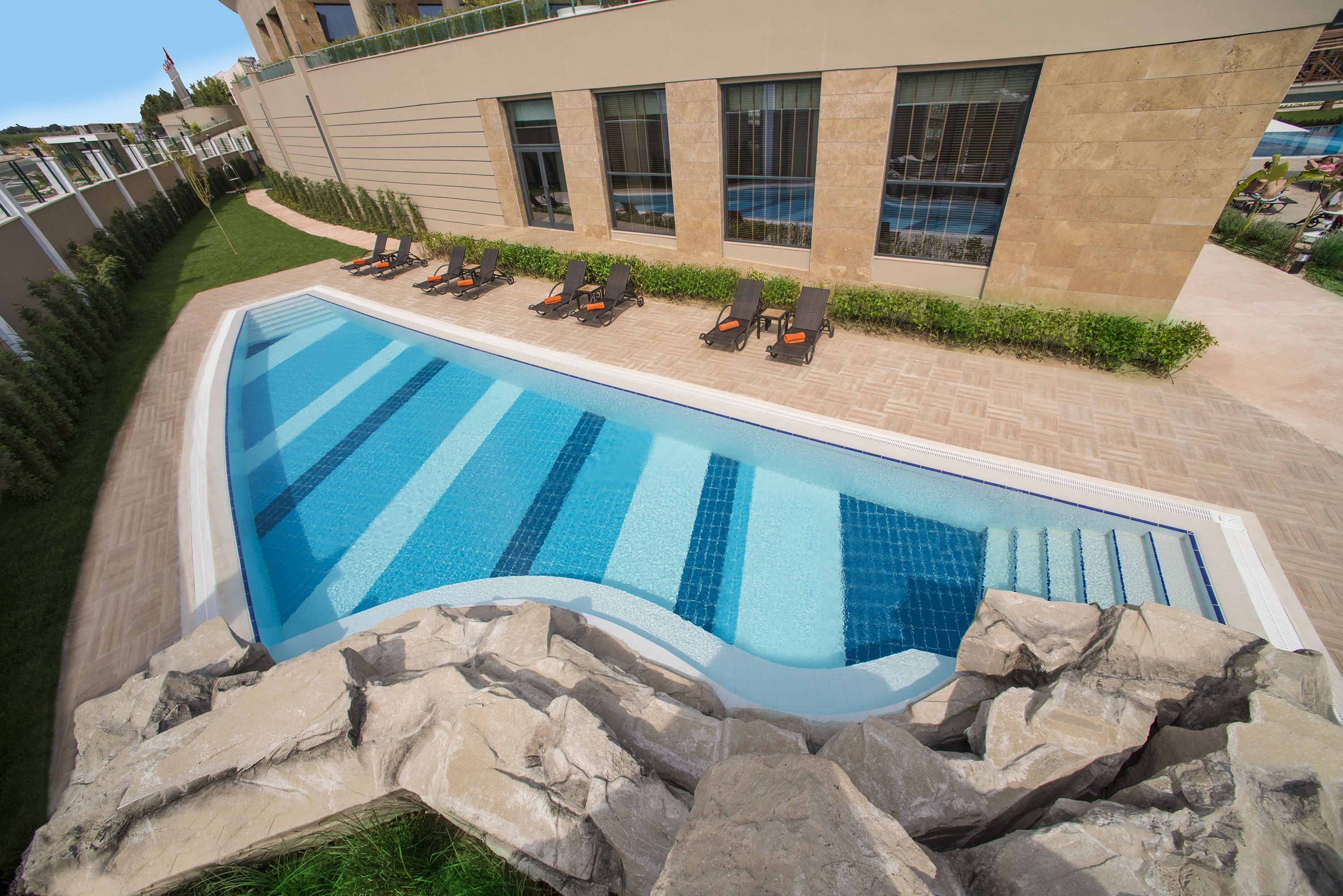 Piscina com espreguiçadeiras, parede de pedra e um edifício com grandes janelas.