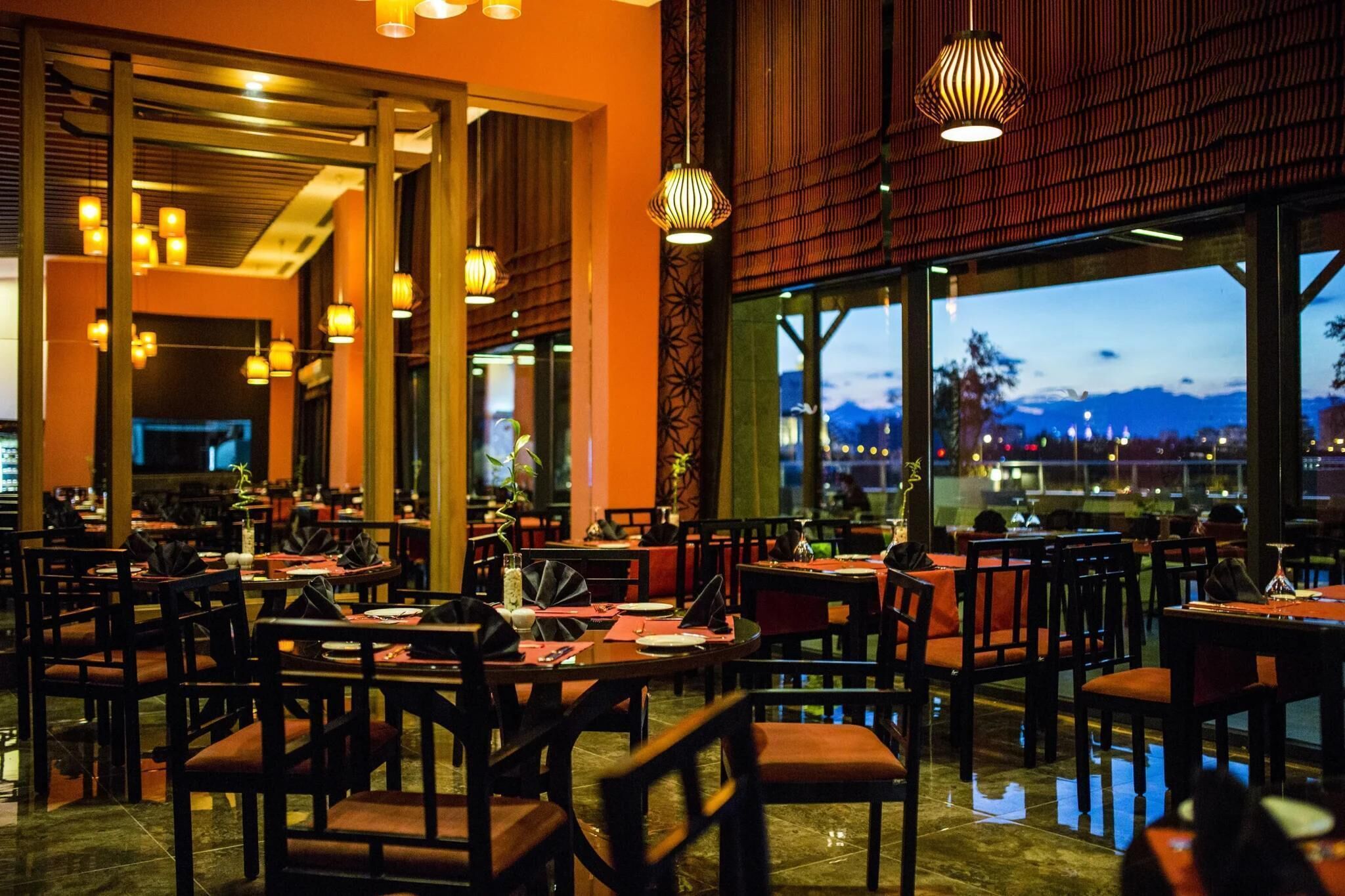 A restaurant with multiple tables and chairs, large windows, and hanging pendant lights.