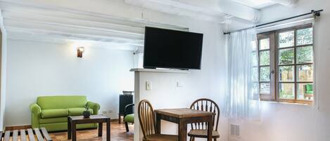 Una habitación con sofá verde, mesa de comedor de madera, sillas, televisor montado en la pared y ventana con cortinas.