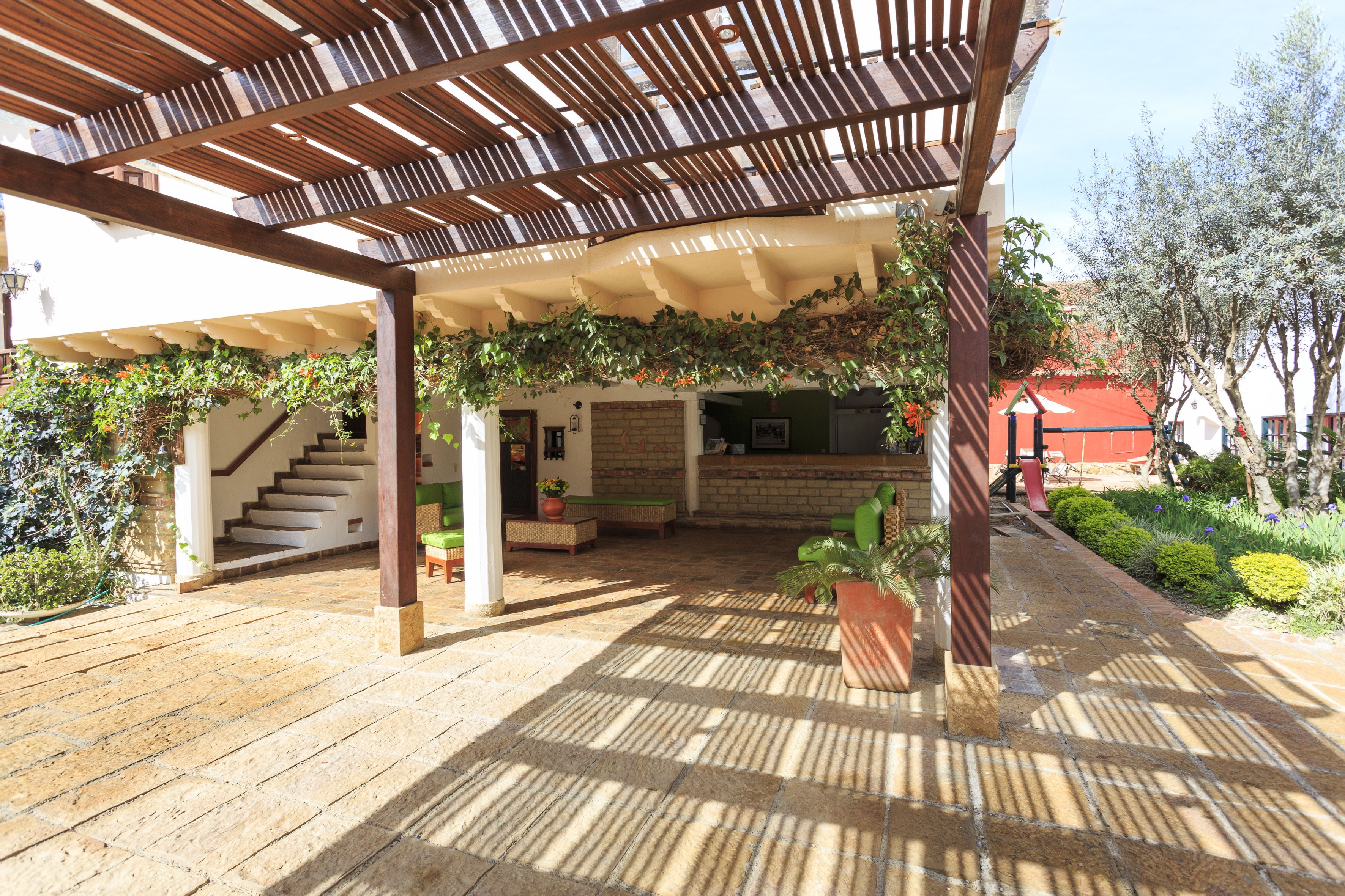 Et overdækket udendørsområde med et træbeklædt pergola, stenbelagte gulve og en trappe op til en etage.