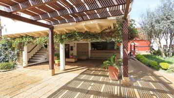 Et overdækket udendørsområde med et træbeklædt pergola, stenbelagte gulve og en trappe op til en etage.