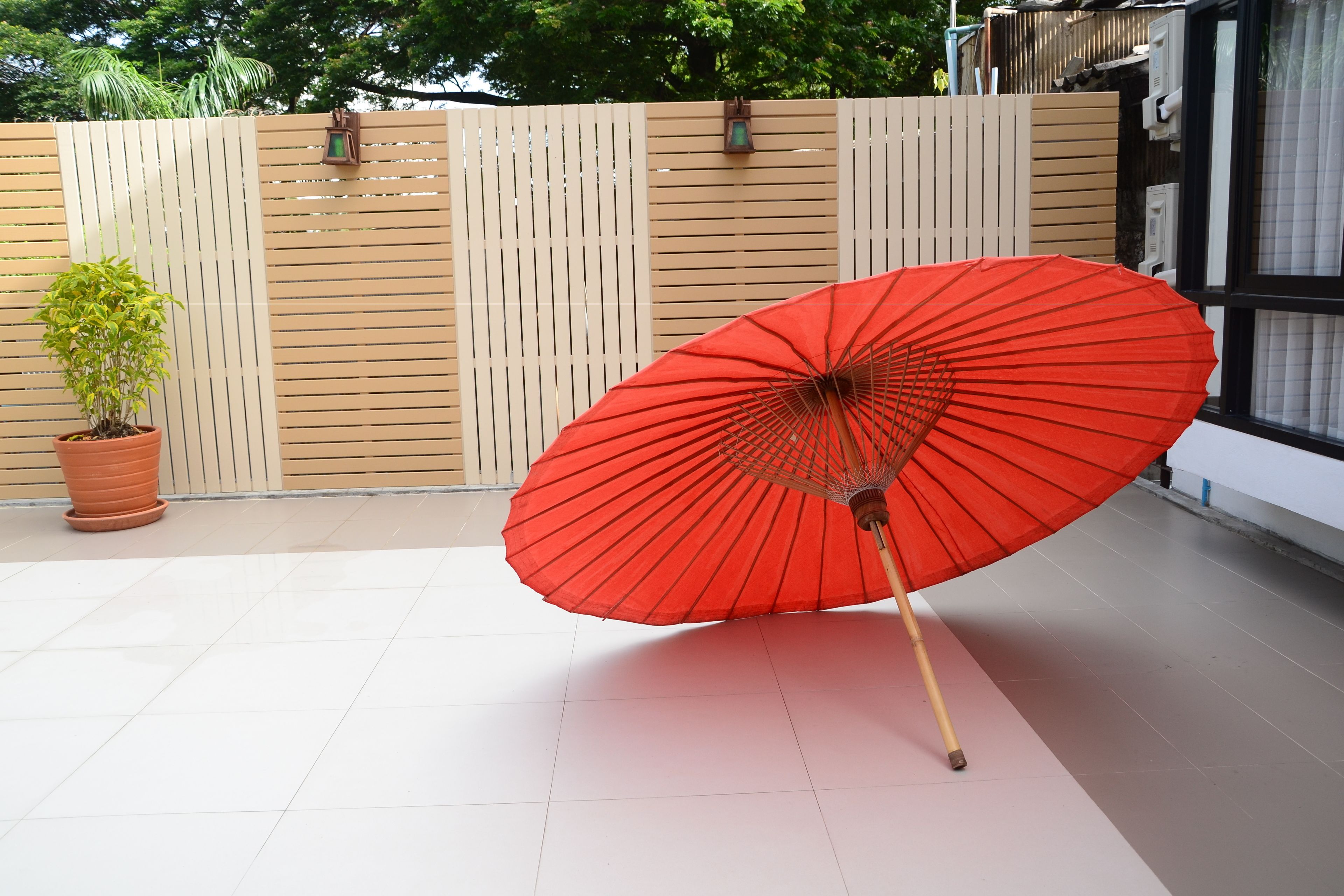 Un parapluie rouge sur un sol carrelé, une plante en pot et une clôture en bois.