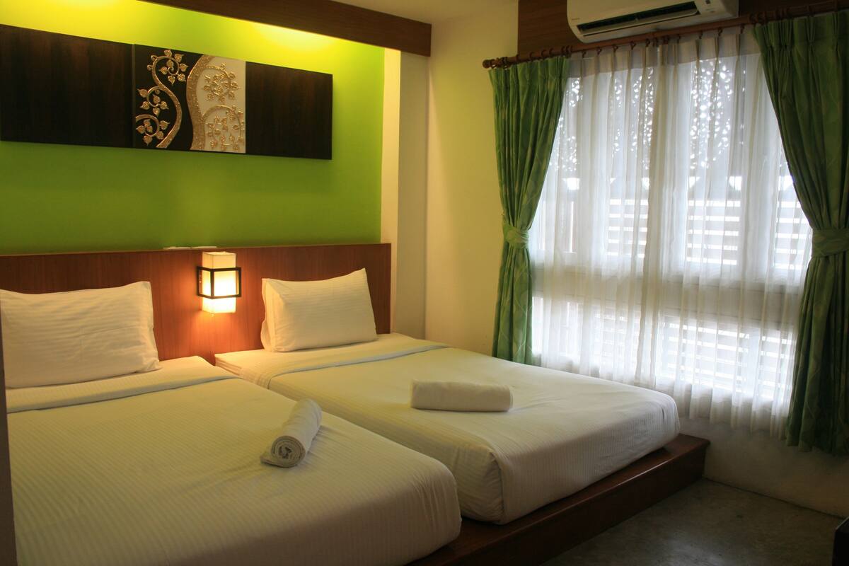 A hotel room with two beds, a wall-mounted air conditioner, a decorative wall piece, and green curtains.