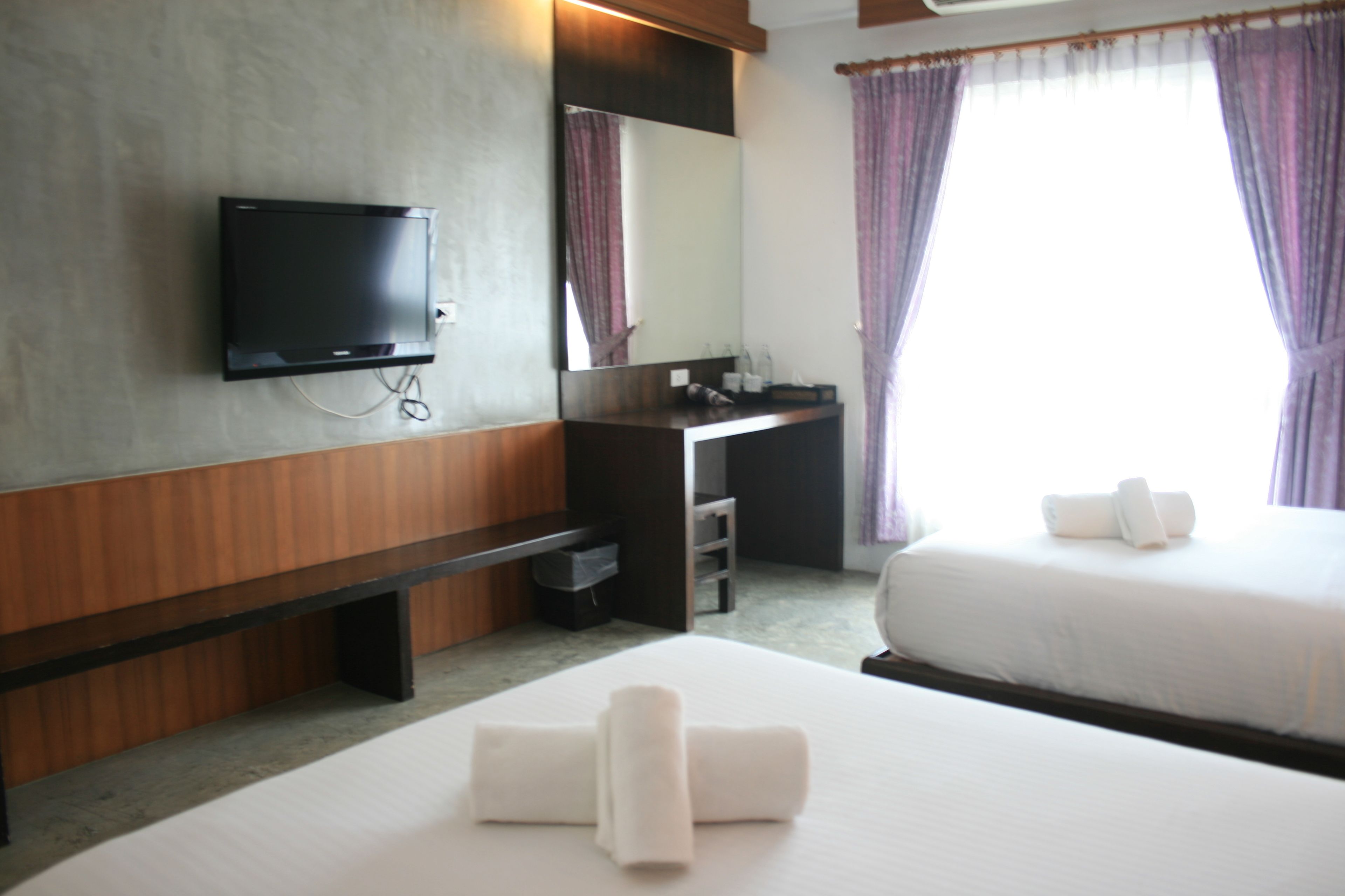 A hotel room with a flat-screen TV, a desk with a mirror, a bed with white linens, and a window with purple curtains.