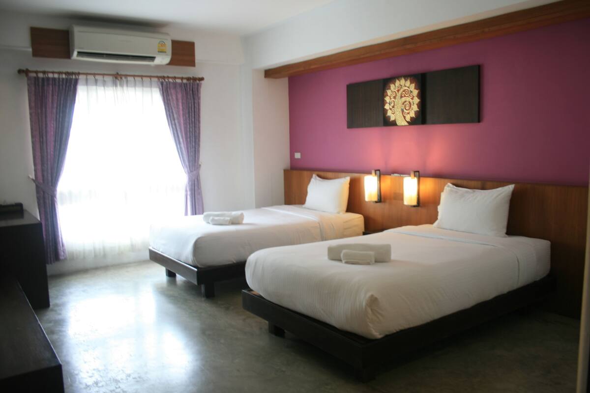 A hotel room with two beds, a large window with curtains, an air conditioning unit, and a wall-mounted light fixture.