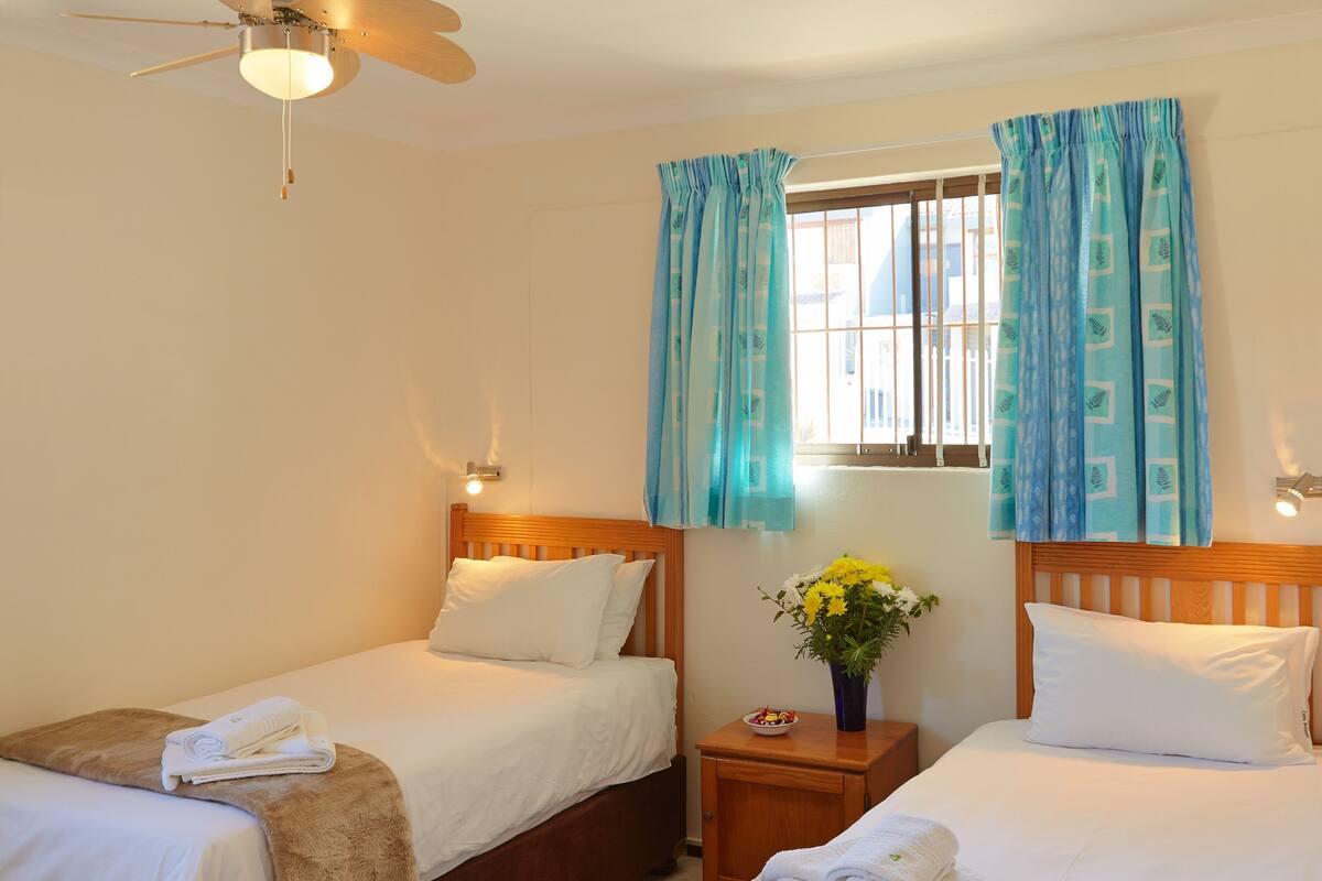 Ein Hotelzimmer mit zwei Betten, einem Deckenventilator, einem Fenster mit Vorhängen, einem Nachttisch mit Blumenvasen und einem blaugrünen Teppich.