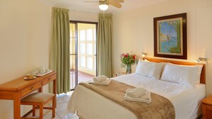 A hotel room with a bed, a desk, a chair, a nightstand, a painting, and a window with curtains. - First Group Costa Smeralda (Margate)