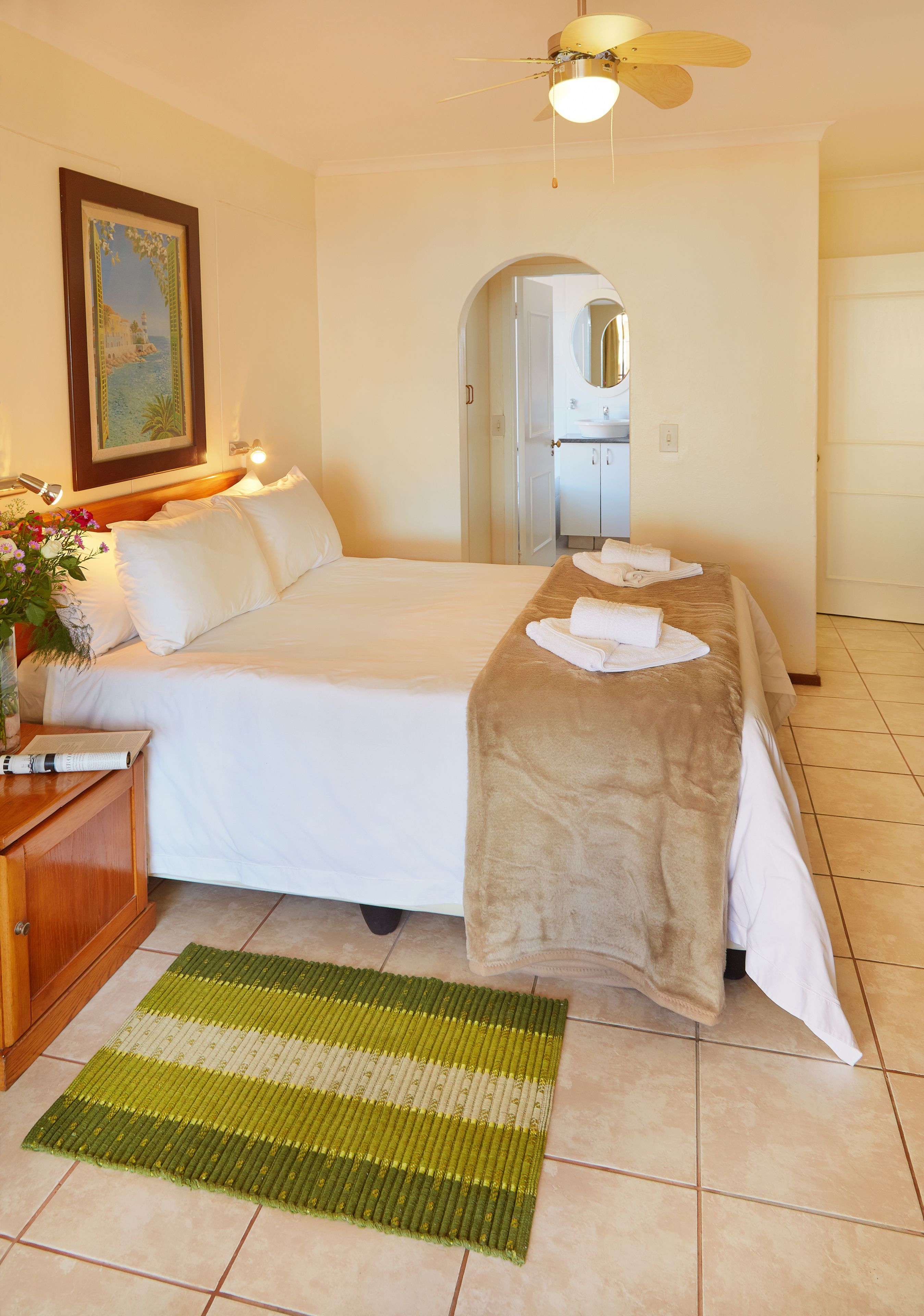 Une chambre d’hôtel avec un lit, une table de chevet en bois, un tapis à rayures vertes et blanches, un ventilateur de plafond et un tableau accroché au mur.