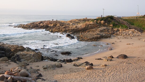 A rocky beach with a staircase leading down to the shore, a sandy beach, and waves crashing against the rocks. - First Group Costa Smeralda (Margate)