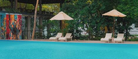 A swimming pool with lounge chairs and umbrellas, surrounded by greenery and a building with a colorful mural.