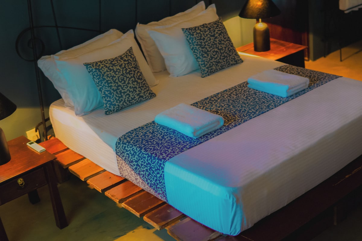 Una cama bien hecha con ropa de cama y almohadas estampadas, una mesita de noche con lámpara y un banco de madera.