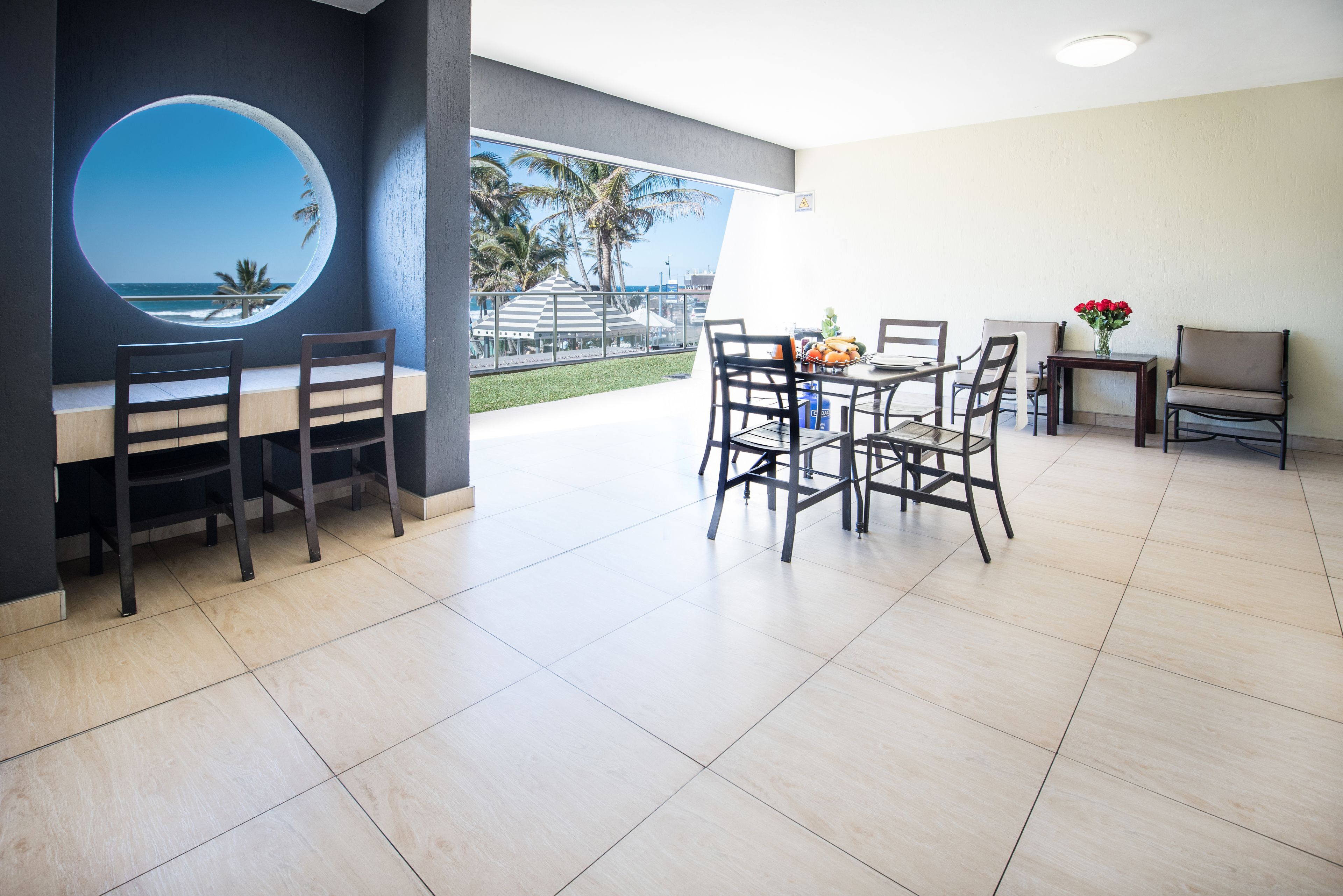 A modern dining area with a round window overlooking a beach, a table with chairs, and a small table with flowers.