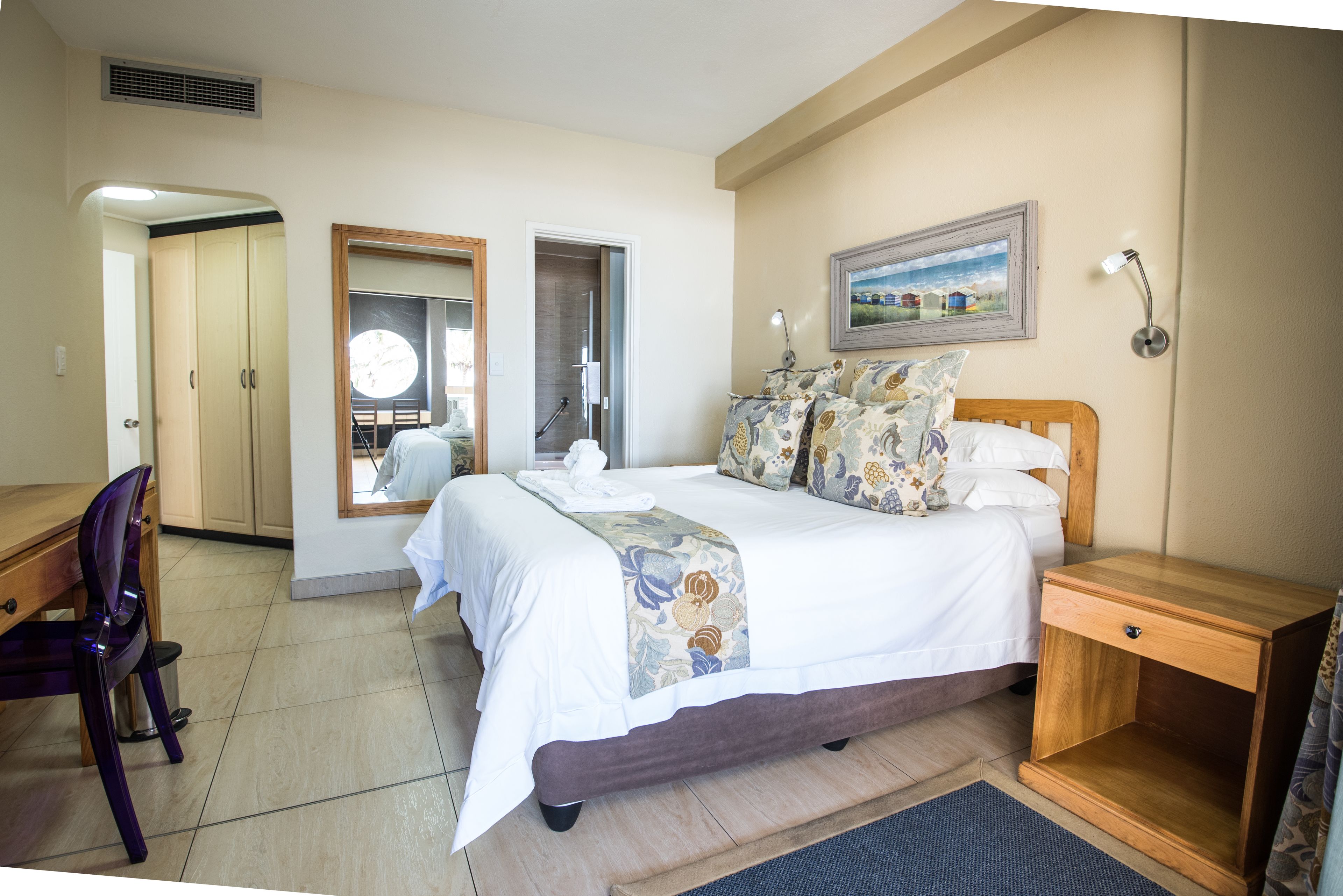 A hotel room with a bed, a wooden nightstand, a desk with a chair, a mirror, and a picture on the wall.