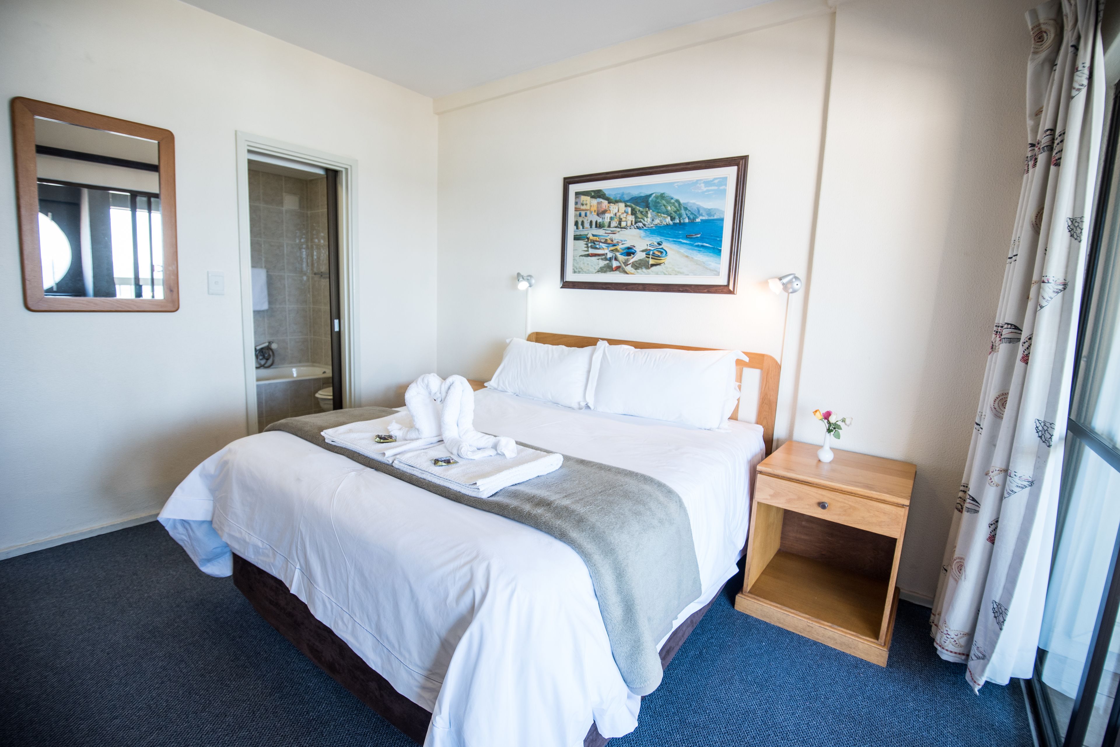 A hotel room with a bed, a nightstand, a painting, and a bathroom door.