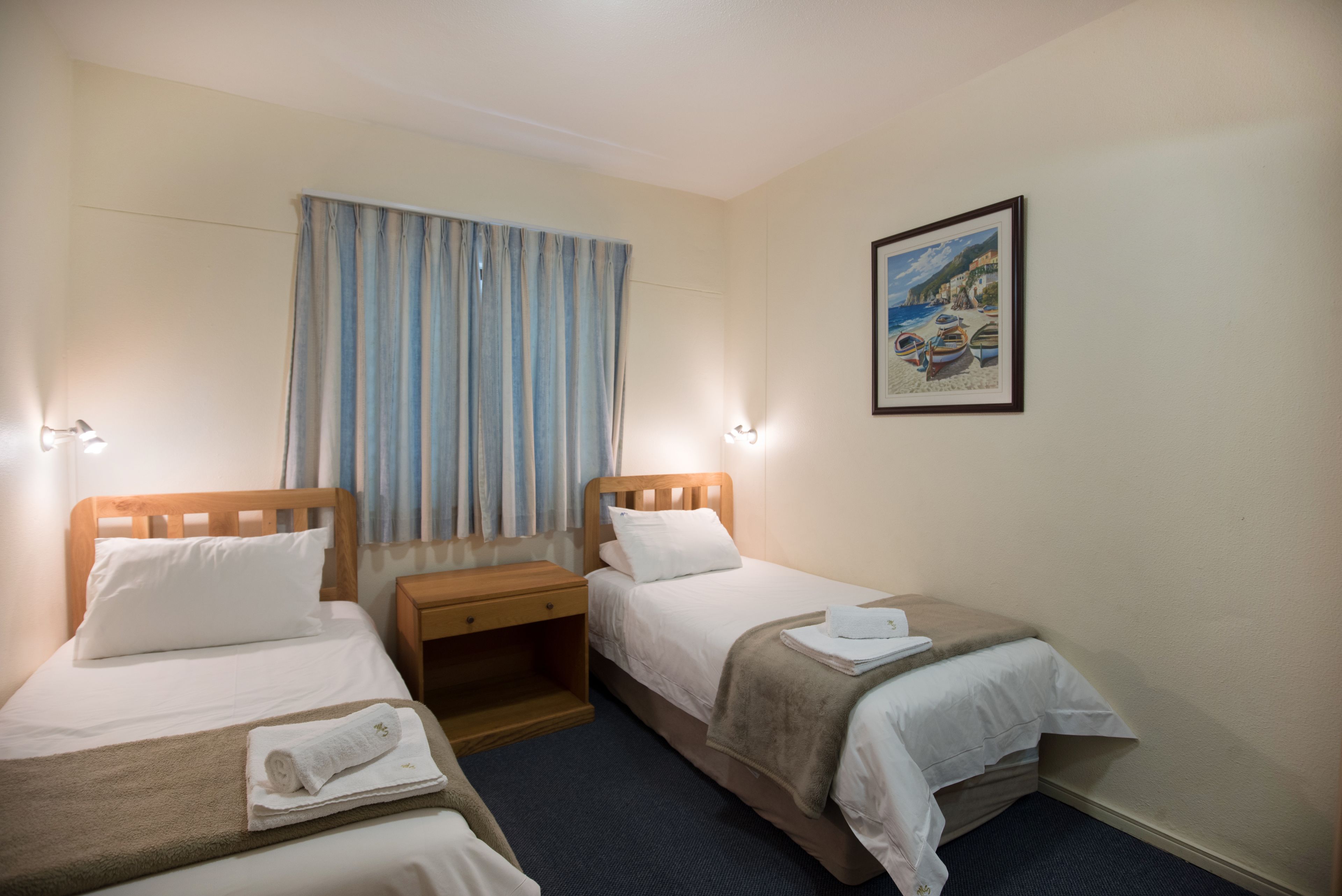 A hotel room with two single beds, a wooden nightstand, a small table, and a painting on the wall.