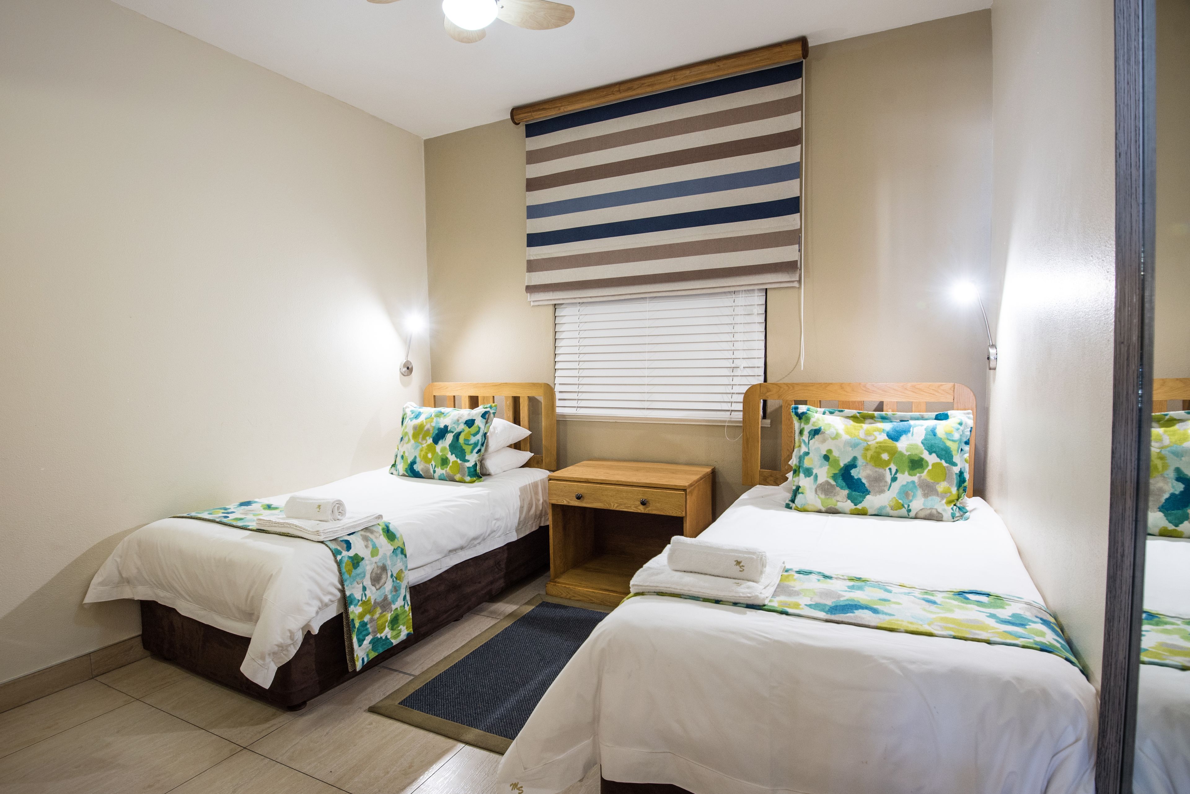 Une chambre d’hôtel avec deux lits simples, une petite table de chevet en bois, un store à rayures et un ventilateur de plafond.