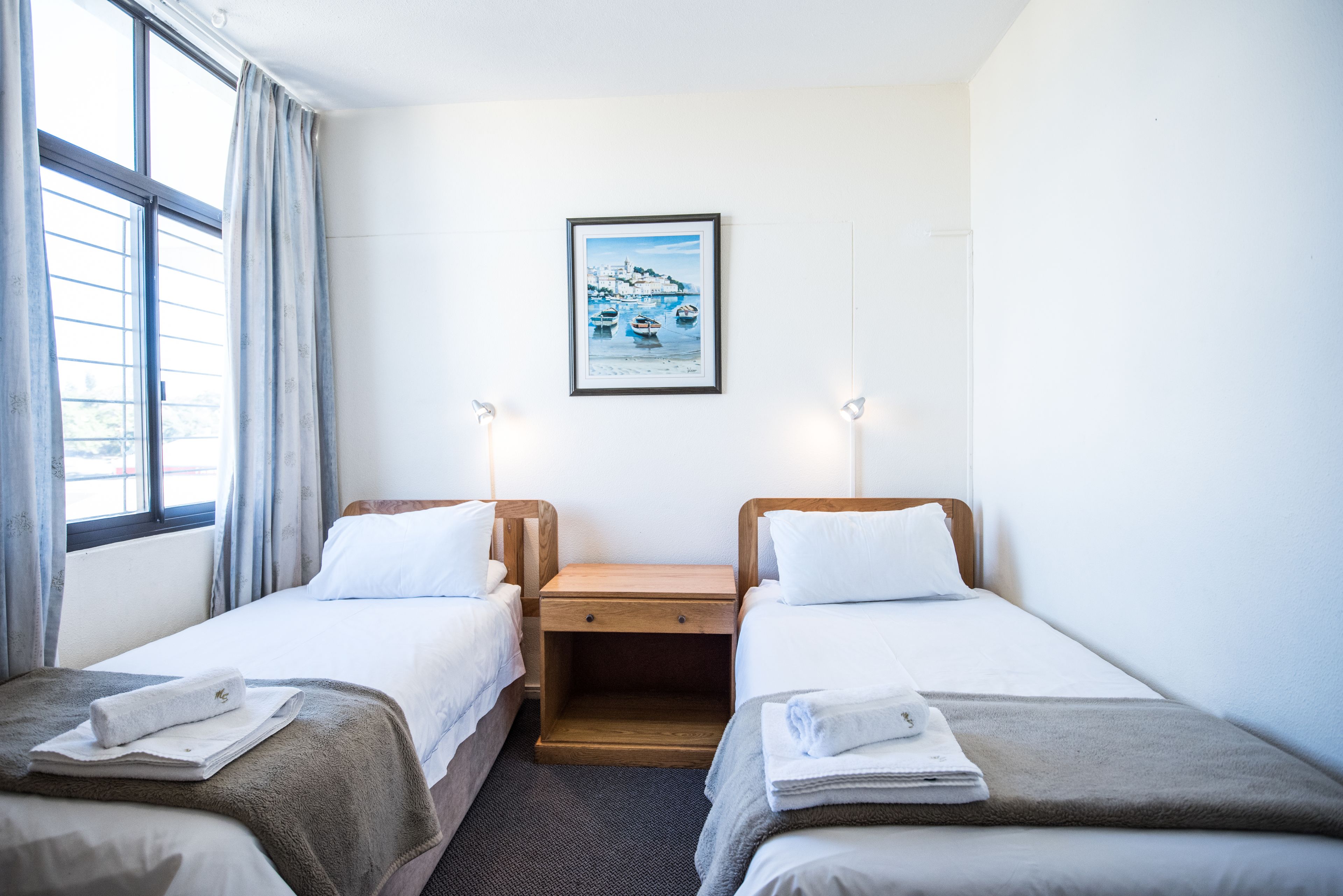 Una habitación de hotel con dos camas individuales, una mesita de noche, un cuadro en la pared y una ventana con cortinas.