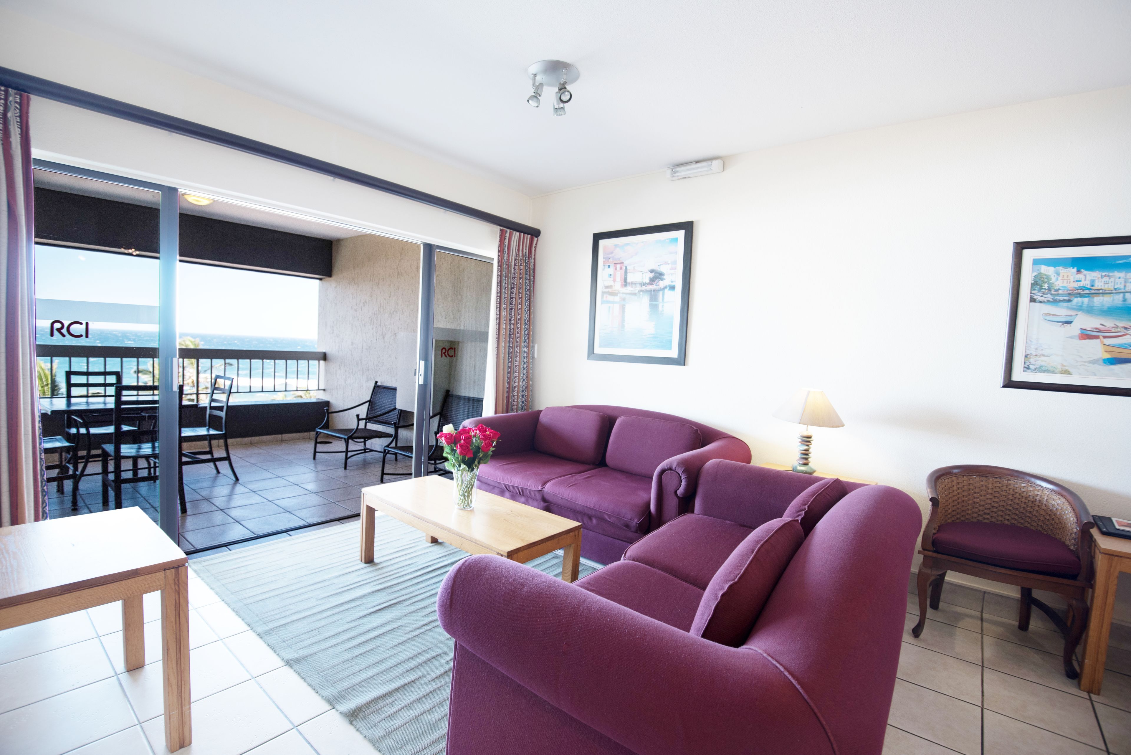 Una sala de estar con un sofá púrpura, una mesa de centro de madera y una silla. Hay un balcón con vista al mar y una mesa con sillas afuera.