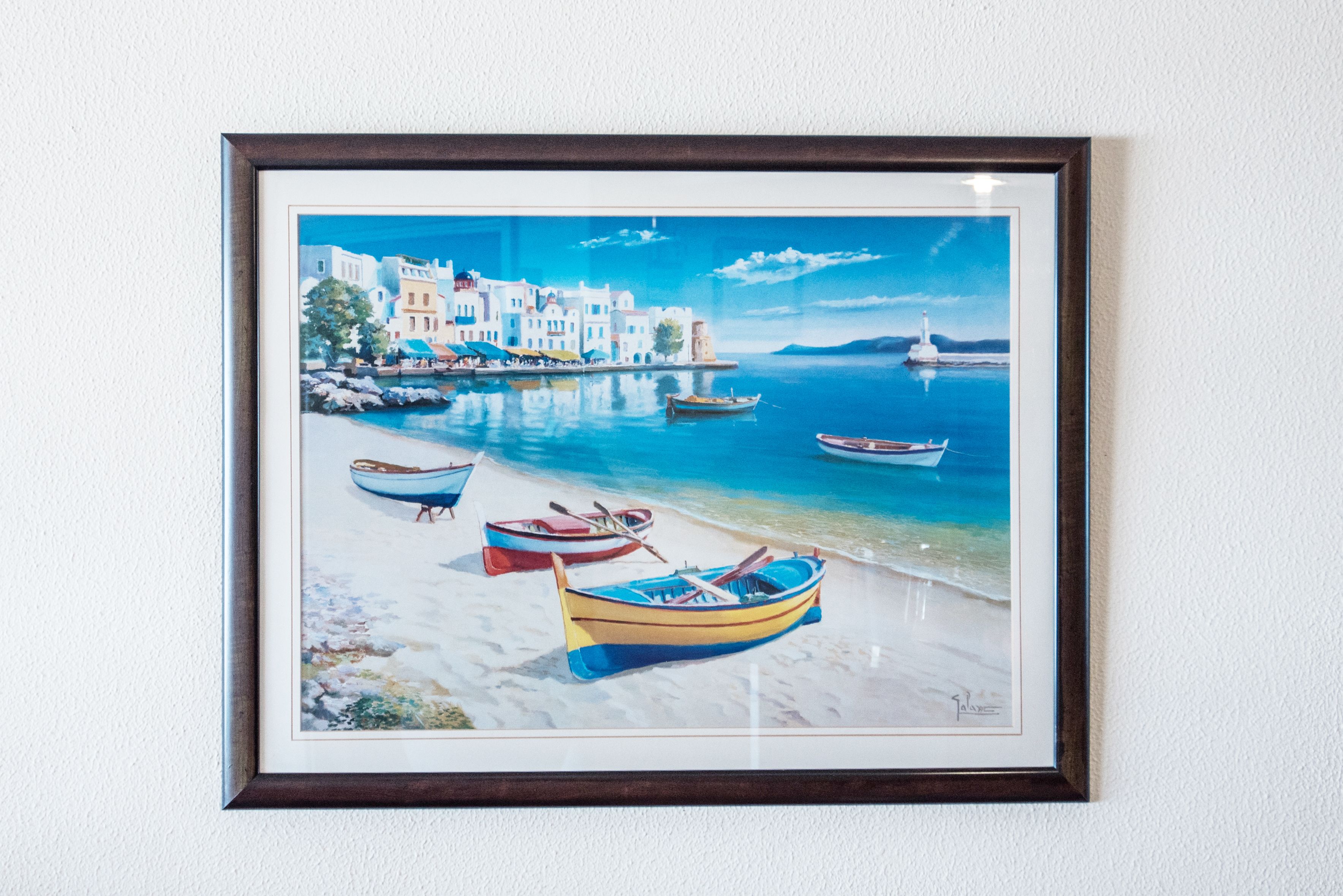 A framed painting of a coastal scene with boats, buildings, and a lighthouse.