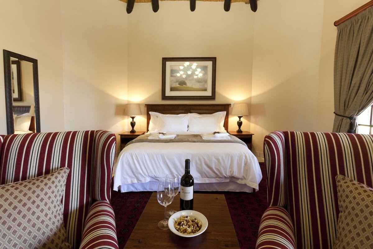Une chambre d’hôtel avec un grand lit, deux tables de chevet avec des lampes, une tête de lit en bois, un tableau encadré au-dessus du lit, et une table à manger avec une bouteille de vin et un bol de noix.