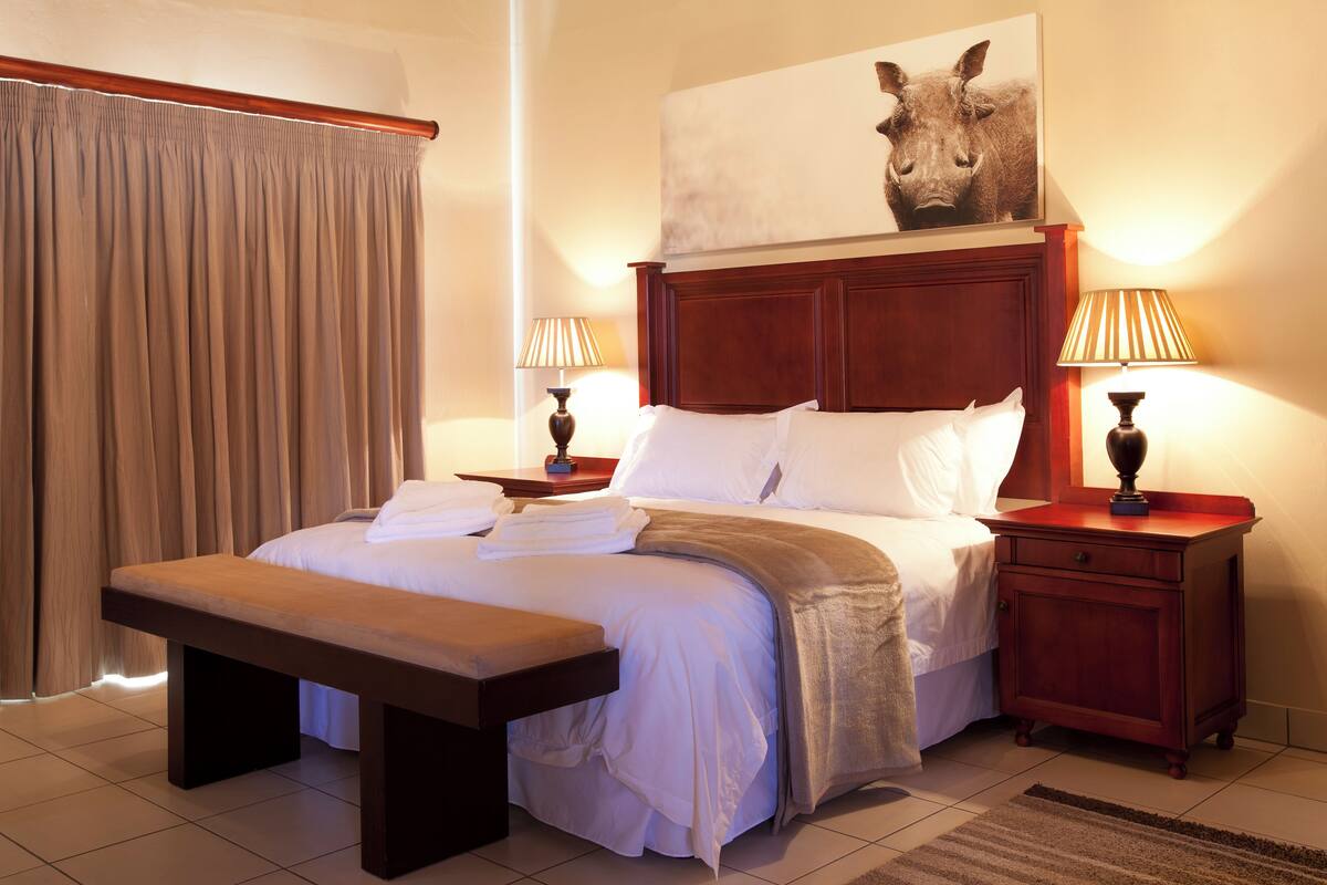 Une chambre d’hôtel avec un grand lit, deux tables de chevet avec des lampes, un banc, une tête de lit et un grand tableau au mur.