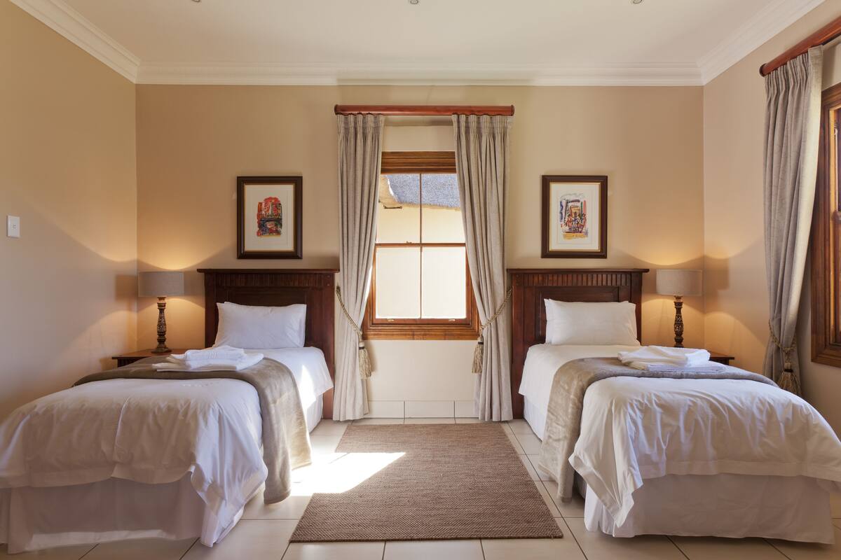 Une chambre d’hôtel avec deux lits, une grande fenêtre avec des rideaux, des œuvres d’art encadrées accrochées aux murs et une tête de lit en bois.