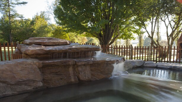 A naturalistic water feature with a cascading waterfall over rocks, surrounded by a wooden fence and trees. - Whispering Waters (Nottingham Road)