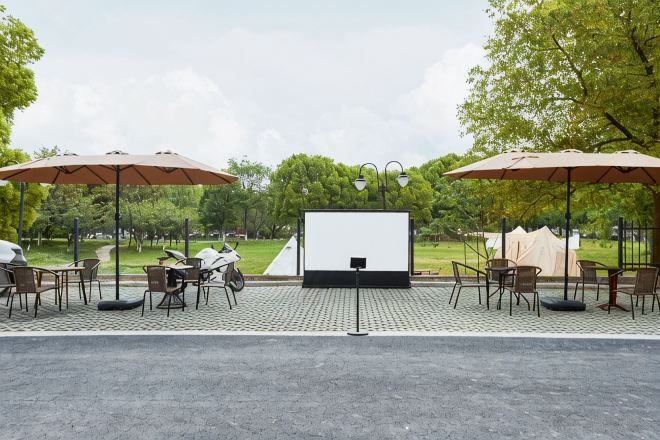Una pantalla de cine al aire libre con sillas y sombrillas, rodeada de árboles.