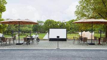 Una pantalla de cine al aire libre con sillas y sombrillas, rodeada de árboles.