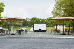 Uma tela de cinema ao ar livre com cadeiras e guarda-sóis, cercada por árvores.