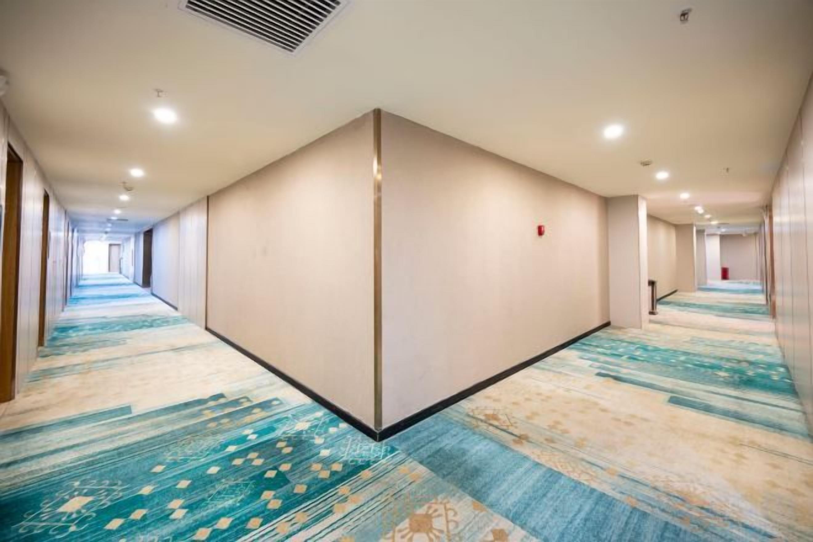 A hotel corridor with carpeted flooring, a central partition, and doors on both sides.