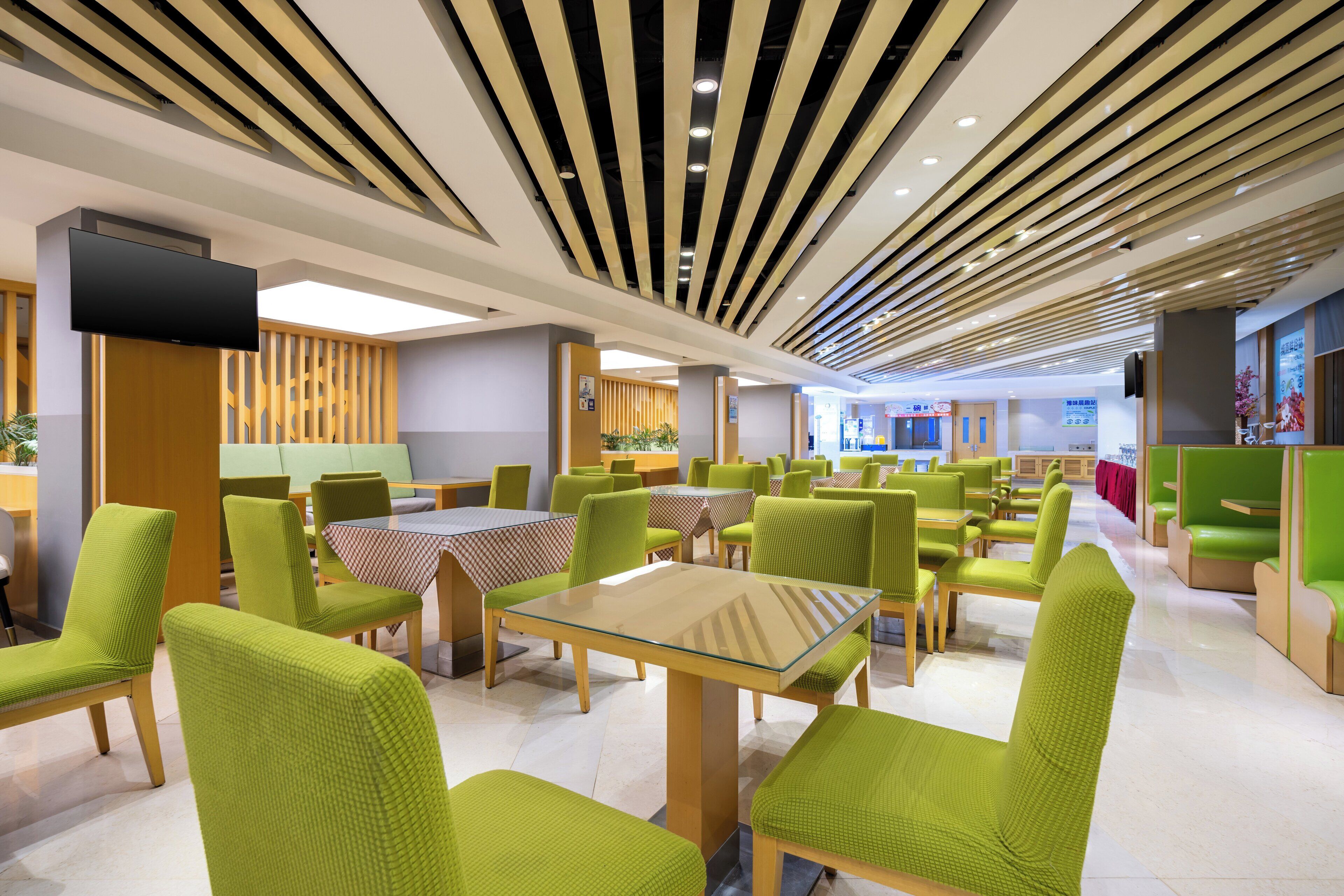A modern dining area with green upholstered chairs and tables, wooden ceiling panels, and a flat-screen TV mounted on the wall.
