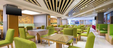 A modern dining area with green upholstered chairs and tables, wooden ceiling panels, and a flat-screen TV mounted on the wall.