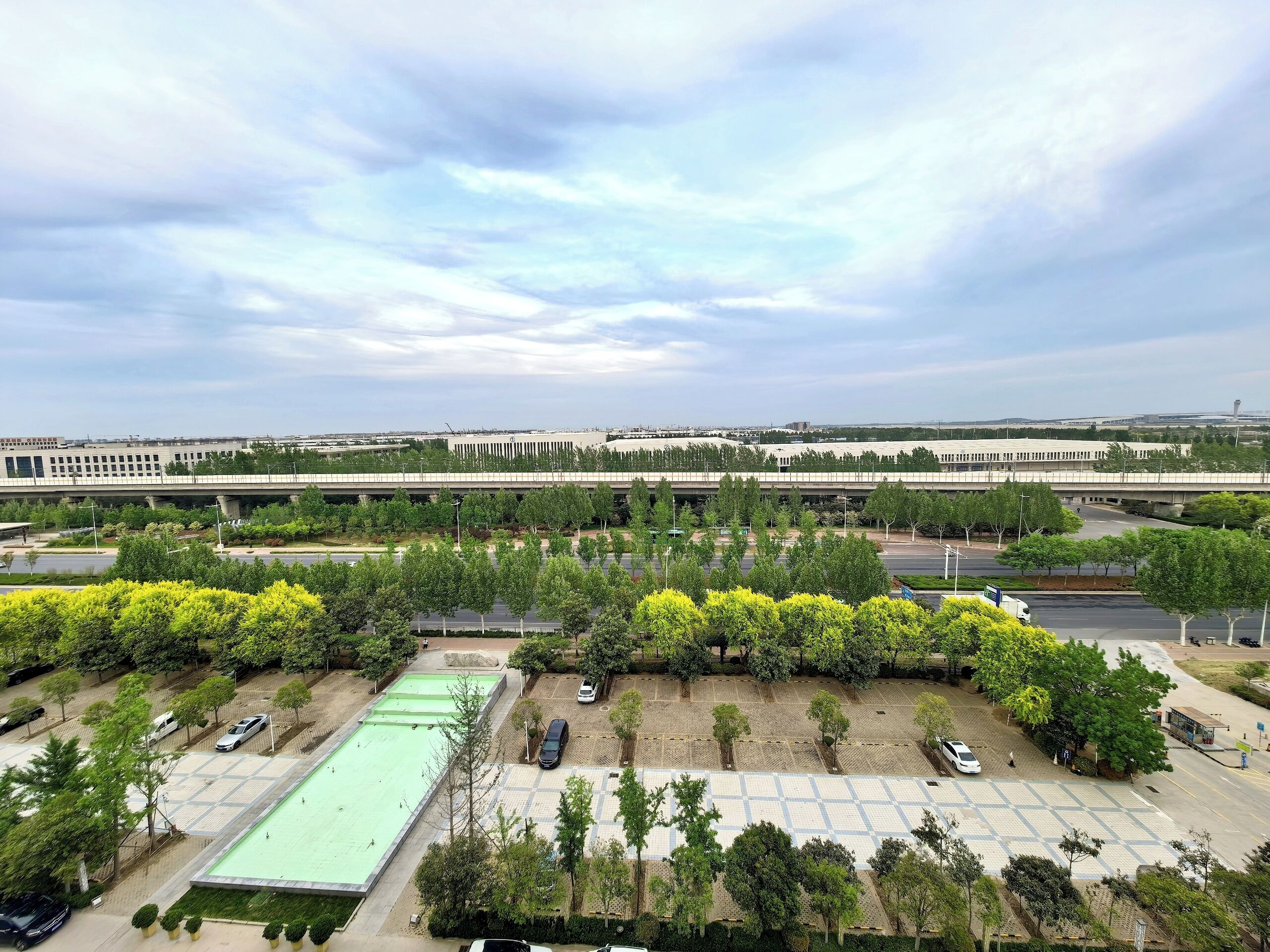 An aerial view of a parking lot with multiple rows of parked cars, surrounded by trees and greenery.