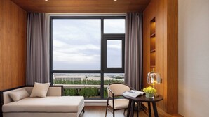Een moderne hotelkamer met een groot raam, een bank, een stoel, een kleine tafel met een lamp en uitzicht op een stadsbeeld.