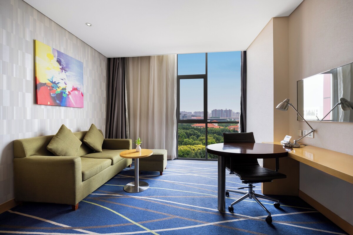 Une chambre d’hôtel avec un canapé, une petite table, un bureau avec une chaise, une grande fenêtre donnant sur la ville et une peinture abstraite aux couleurs vives accrochée au mur.