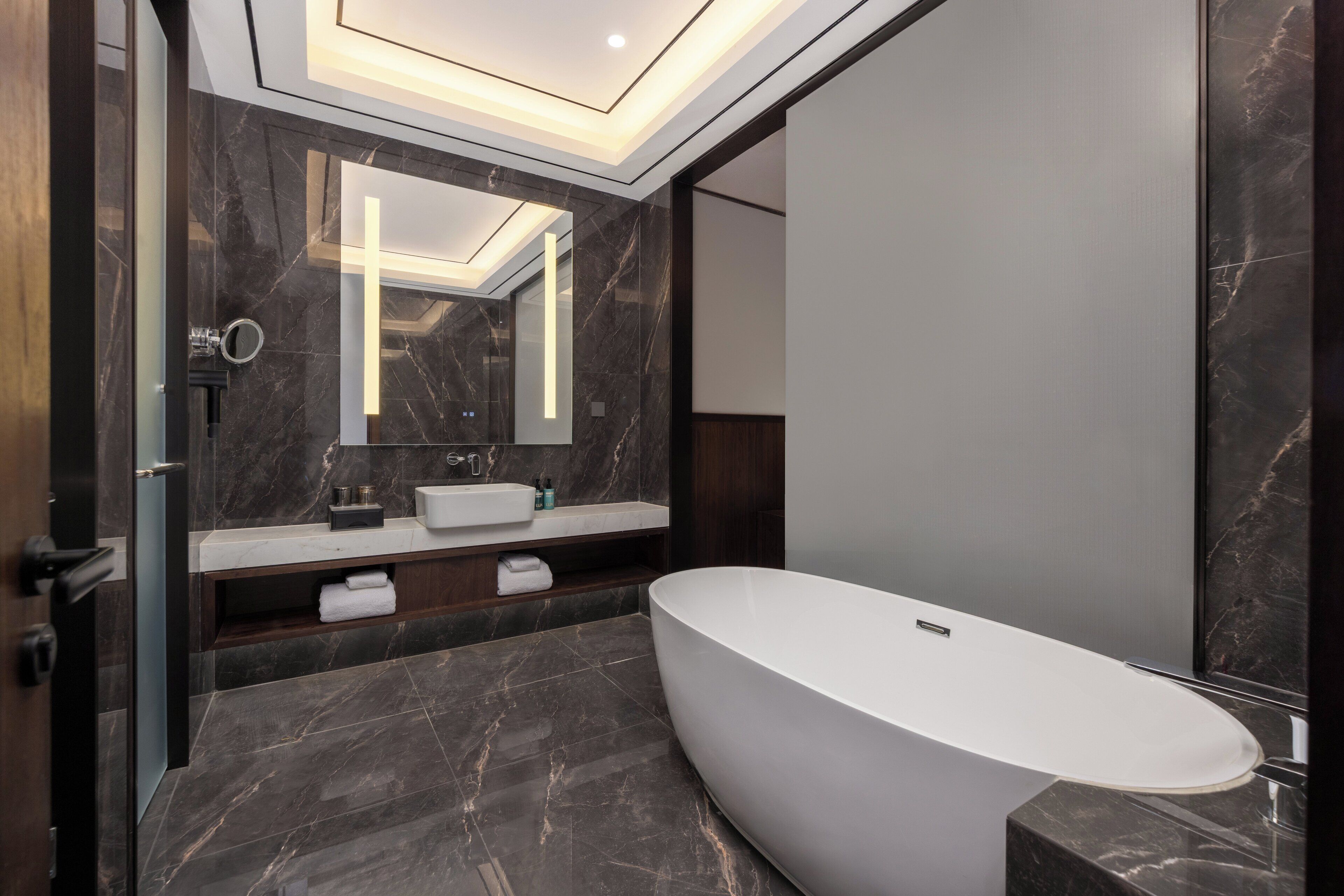 Un baño moderno con una bañera independiente, suelo de mármol oscuro y un espejo grande sobre el lavabo.