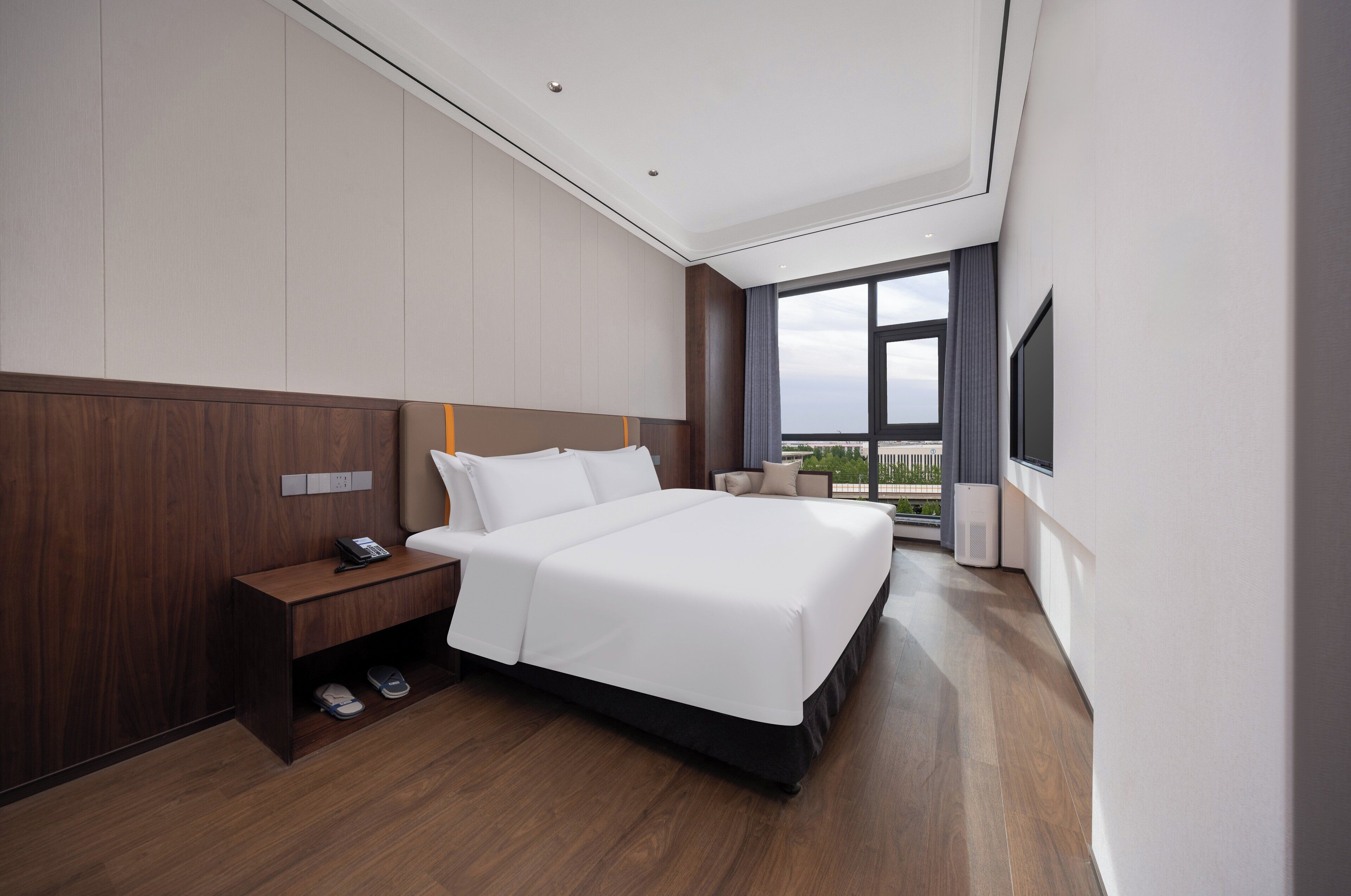 A modern hotel room with a large bed, a bedside table with a phone, a shoe rack, and a large window offering a view of the city.