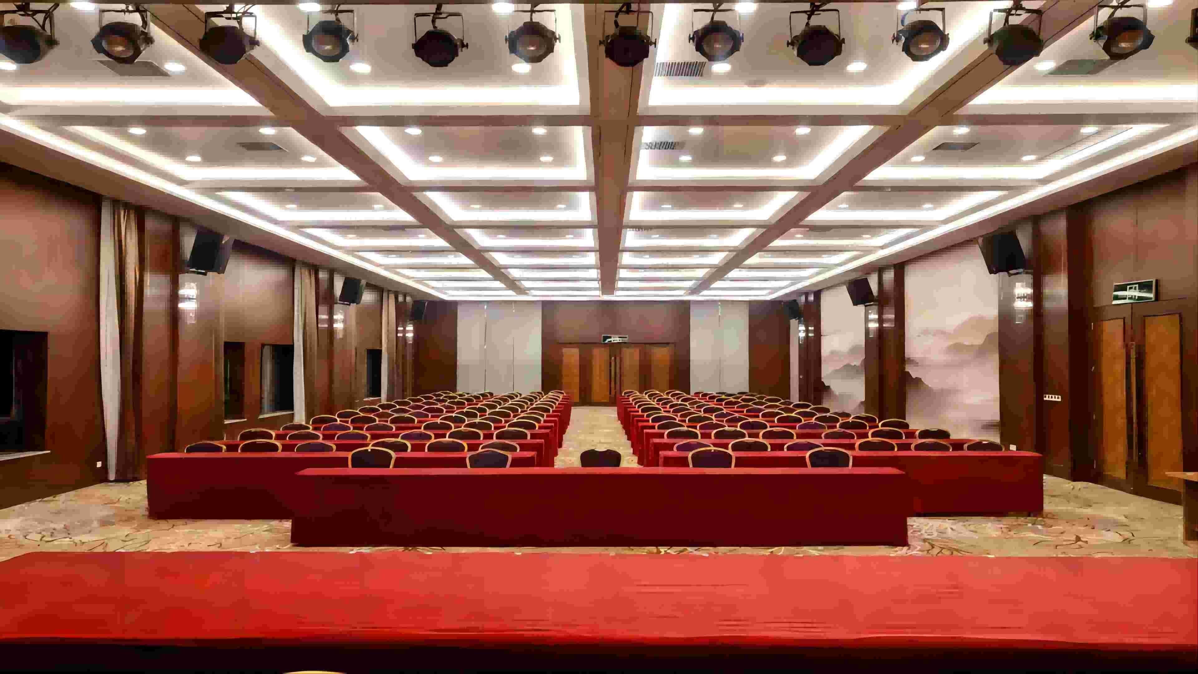 A conference room with rows of red chairs, a stage, and a ceiling with multiple lights.