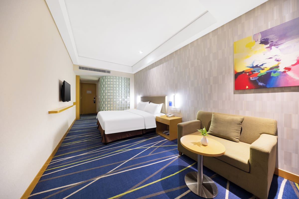Ett hotellrum med en säng, en soffa, ett litet bord med en växt, en tv och en färgstark, abstrakt målning på väggen.