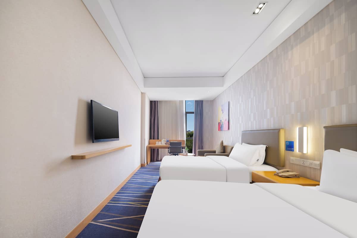 Ett hotellrum med två sängar, en TV, ett skrivbord med en stol och ett fönster med gardiner.